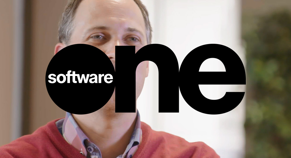 A man in a red sweater is standing in front of the software one logo.