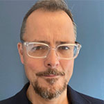 A man with glasses and a beard standing in front of a blue wall.