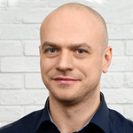 A bald man in a blue shirt standing in front of a brick wall.